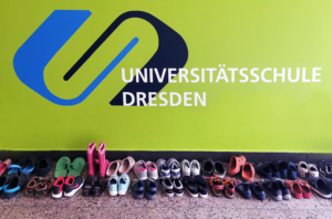 Vor einer grünen Wand steht eine Reihe vieler bunter Schuhe. An der Wand ist das Logo und der Schriftzug Universitätsschule Dresden aufgebracht.