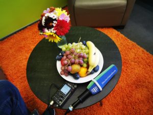Auf dem Couchtisch im Schulleiterinnenbüro der Universitätsschule liegen das Deutschlandfunk-Mikrofon und ein Aufnahmegerät neben einer Schale mit Früchten und einem bunten Blumenstrauß. Der Tisch steht auf einem orangefarbenen Teppich vor einer leuchtend grünen Wand.